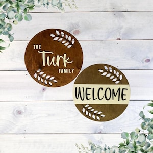 May include: Two wooden signs with a brown background and white lettering. The first sign says "The Turk Family" with a leafy design around the text. The second sign says "Welcome" with a similar leafy design.