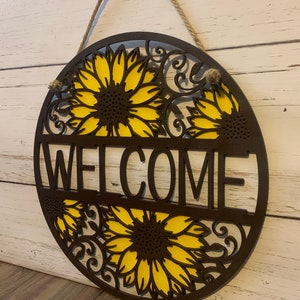 May include: A round wooden welcome sign with a sunflower design. The sign is painted brown and yellow and has the word "WELCOME" in the center.