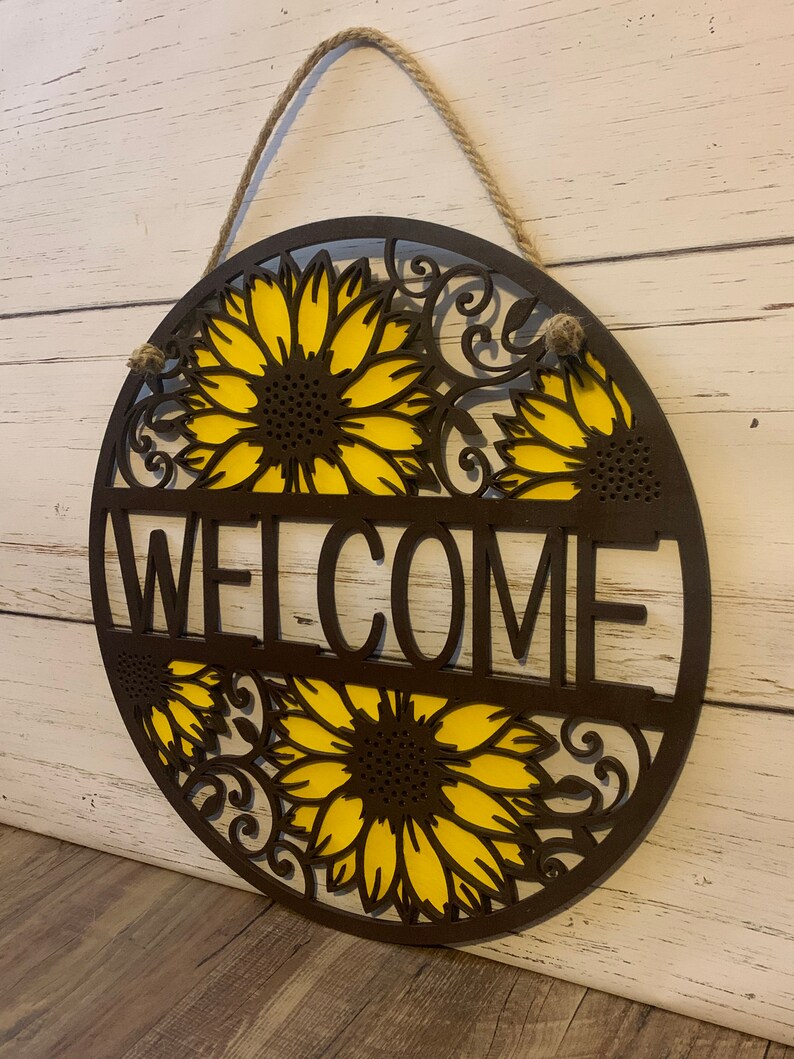 May include: A round wooden welcome sign with a sunflower design. The sign is painted brown and yellow and has the word "WELCOME" in the center.