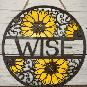 May include: A round wooden sign with a brown background and yellow sunflowers. The word "WISE" is cut out of the wood in the center of the sign.