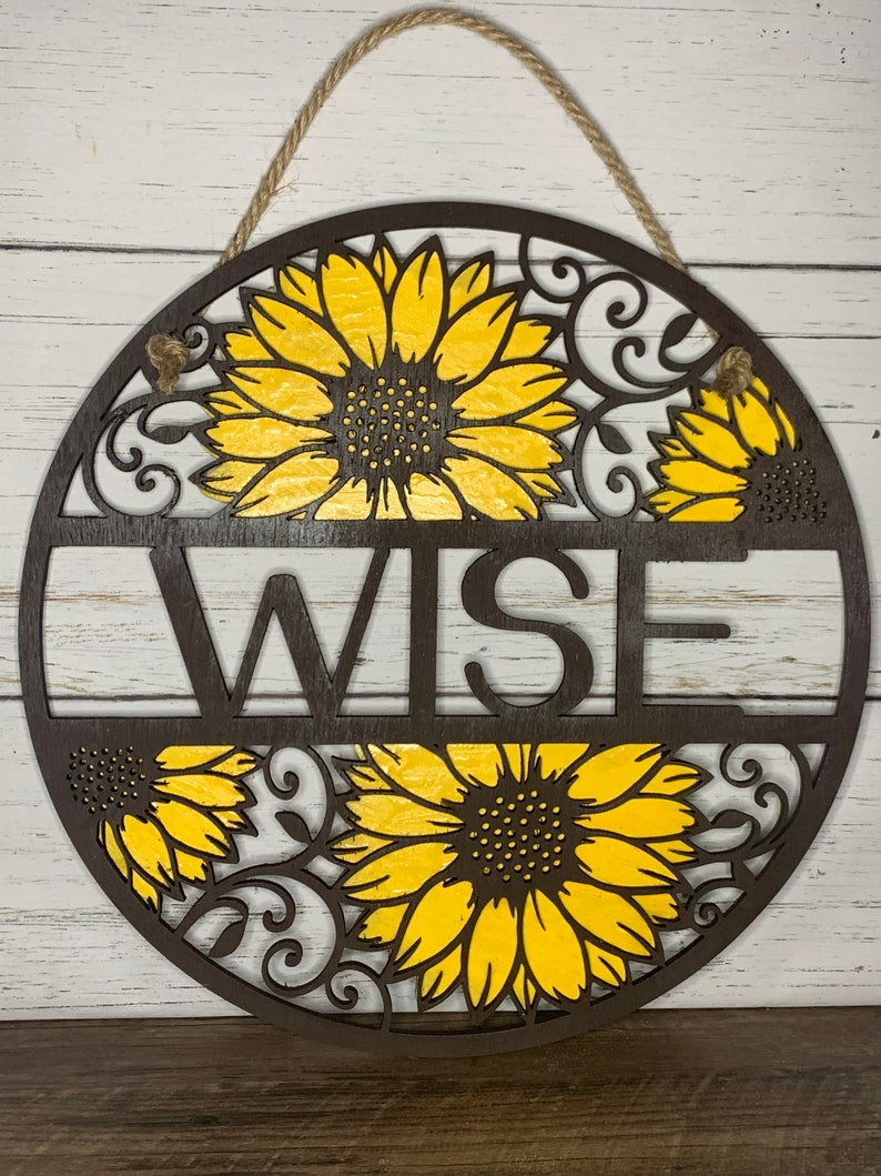 May include: A round wooden sign with a brown background and yellow sunflowers. The word "WISE" is cut out of the wood in the center of the sign.