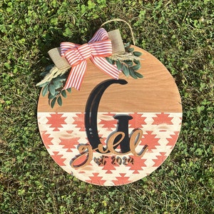 May include: A round wooden sign with a red and white geometric pattern. The sign has a black letter "G" and the text "est 2024" in black. The sign is decorated with a red and white striped bow and green leaves.