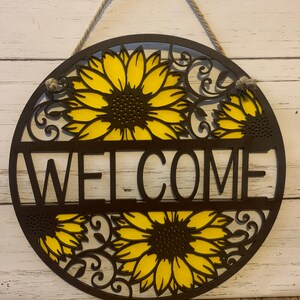 May include: A round wooden welcome sign with a brown background and yellow sunflowers. The sign has a rope hanger and the word "WELCOME" is cut out in the center.