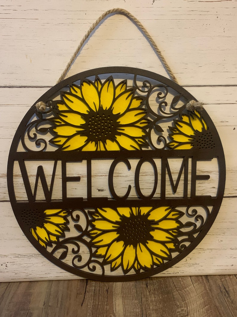 May include: A round wooden welcome sign with a brown background and yellow sunflowers. The sign has a rope hanger and the word "WELCOME" is cut out in the center.