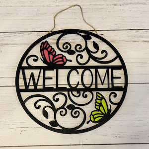 May include: Black metal welcome sign with a decorative floral design and two butterflies, one pink and one green.