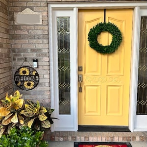 May include: A yellow front door with a green boxwood wreath hanging on it. There is a welcome sign with sunflowers on the brick wall to the left of the door. A welcome mat with the words "love grows here" is on the ground in front of the door.