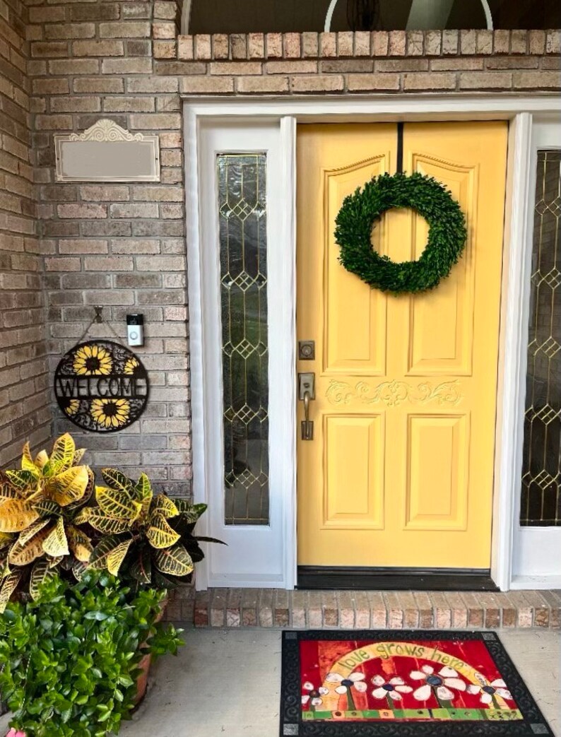 May include: A yellow front door with a green boxwood wreath hanging on it. There is a welcome sign with sunflowers on the brick wall to the left of the door. A welcome mat with the words "love grows here" is on the ground in front of the door.
