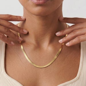 May include: A gold-colored herringbone chain necklace. The necklace is worn around the neck and is held by two hands. The necklace is made of a flat, woven design. The person is wearing a cream-colored ribbed top.