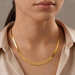 May include: A gold-toned herringbone chain necklace featuring a flat, flexible metal band. The centre of the chain is engraved with the text SOPHIA-MARY-MOM-DAD, flanked by two small heart symbols, creating a personalised piece of jewellery.