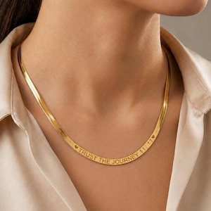 Op de afbeelding: Een goudkleurige visgraatketting met een gegraveerde inscriptie "TRUST THE JOURNEY 11:11" geflankeerd door twee kleine hartjes. De platte, flexibele metalen schakels vormen een gladde, doorlopende band rond de hals.