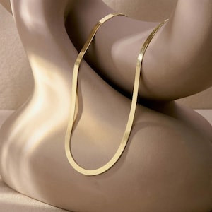 May include: A gold herringbone chain necklace. The necklace is draped over a mannequin. The chain has a flat, woven design, reflecting light. The background is a neutral beige colour.
