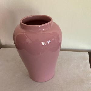Könnte beinhalten: Eine rosafarbene Keramikvase mit rundem Körper und einer breiten Öffnung. Die Vase steht auf einer weißen Tischdecke mit Blumenmuster.