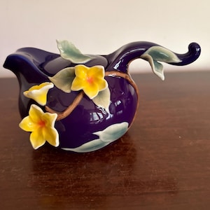May include: A deep blue ceramic creamer featuring three-dimensional yellow flowers with green centers and light green leaves attached to a brown branch. The creamer has a curved handle and a spout, with a glossy finish throughout the piece.