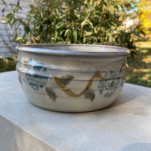 May include: A handmade ceramic bowl with a cream base and a brown rim. The bowl has a decorative band of blue and brown leaf-like patterns around its circumference. The bowl is on a stone surface.
