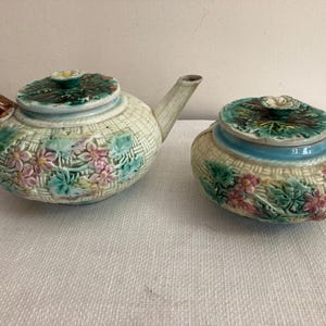 May include: A ceramic teapot and sugar bowl set. The teapot has a brown handle and spout, with a lid featuring a green leaf design. Both pieces are decorated with pink and blue flowers and a woven basket pattern.