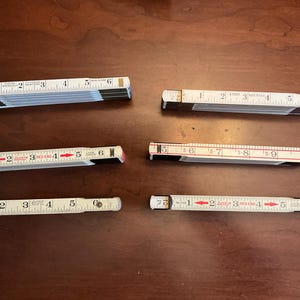 Pode incluir: Seis réguas dobráveis brancas com marcações pretas e vermelhas dispostas sobre uma superfície de madeira castanha. Cada régua é marcada com medidas em polegadas, com números e setas vermelhas. Uma ferramenta de medição clássica.