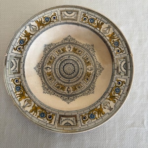May include: A round, cream-colored decorative plate with an ornate design. The plate features a central circular pattern and a border with repeating geometric and floral motifs in gold, blue, and black.