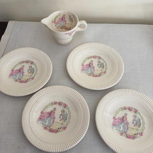 Peut inclure: Un service de vaisselle en porcelaine crème avec un motif décoratif d'un couple en costume d'époque entouré d'une couronne florale. L'ensemble comprend quatre assiettes et une crémière. Les assiettes ont un bord strié.