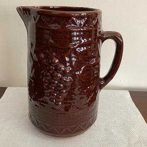 May include: A brown ceramic pitcher with a handle and spout. The pitcher features a raised relief design of grape vines and a textured surface. The pitcher is sitting on a white cloth.