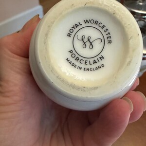 May include: The underside of a white porcelain vessel featuring a black circular stamp that reads Royal Worcester Porcelain Made in England with a central double S logo. The base has a smooth, off-white finish with some visible surface wear.