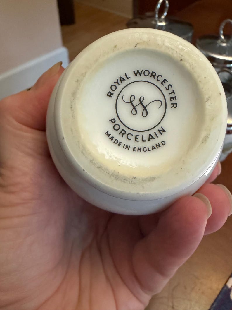 May include: The underside of a white porcelain vessel featuring a black circular stamp that reads Royal Worcester Porcelain Made in England with a central double S logo. The base has a smooth, off-white finish with some visible surface wear.
