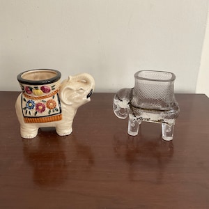 May include: Two ceramic elephant planters, one white with colorful floral designs and the other clear glass with a textured surface.