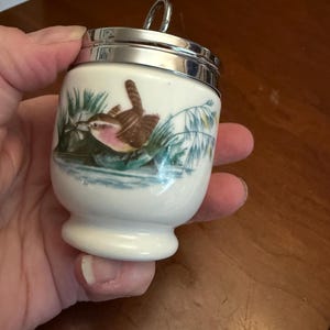 May include: A white ceramic egg coddler with a polished silver-tone metal lid and a small ring handle. The body features a colorful illustration of a small brown bird perched among green reeds and blue water, painted in a detailed, realistic style.
