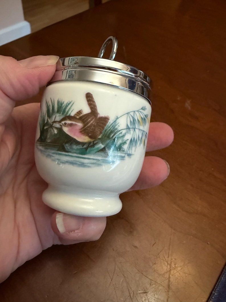 May include: A white ceramic egg coddler with a polished silver-tone metal lid and a small ring handle. The body features a colorful illustration of a small brown bird perched among green reeds and blue water, painted in a detailed, realistic style.