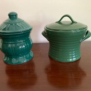 May include: Two green ceramic canisters with lids. The canister on the left has a rounded, fluted design. The canister on the right has a simple, rounded design with two handles.