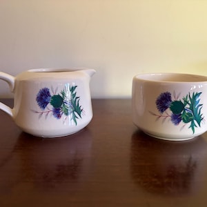 May include: A cream-colored ceramic creamer and sugar bowl set. Both pieces feature a painted graphic of a purple and green thistle flower. The creamer has a handle and a pouring spout, while the sugar bowl is a simple open-top cylindrical vessel.