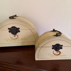 May include: Two off-white, half-circle wooden boxes with decorative metal hardware. Each box features a painted seashell design and a dark brown clasp. The boxes are of different sizes and sit on a dark wooden surface.