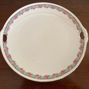 Op de afbeelding: Een ronde, witte serveerschaal met een rand van roze rozen en een lichtblauw traliewerkpatroon. De schaal heeft twee handvatten en een gouden rand. Een vintage stuk voor het serveren van eten.