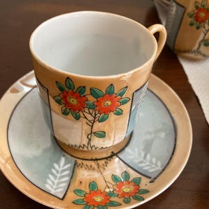 May include: A vintage teacup and saucer set. The cup and saucer are decorated with orange flowers and green leaves. The set has a light orange and blue color scheme. The cup has a small handle. Additional cups and saucers are visible in the background.