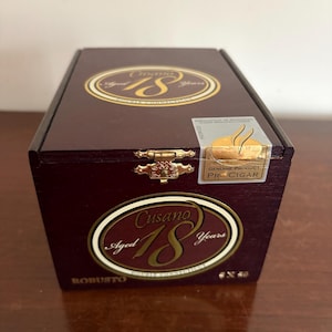Può includere: Una scatola rettangolare in legno marrone scuro con coperchio incernierato. La scatola presenta etichette ovali dorate con la scritta "Cusano Aged 18 Years". La scatola è etichettata "Robusto" e "Genuine Product PR CIGAR".