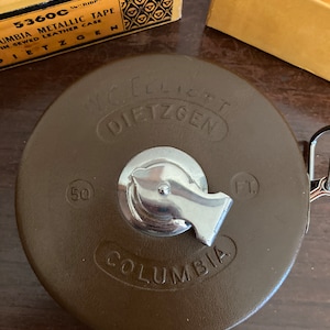 May include: A brown leather measuring tape with a silver metal case. The tape is labeled "Columbia" and "W. & L. E. Dietzgen" in black lettering. The tape is marked with "50" and "Ft" in white lettering.
