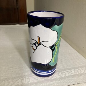 May include: A cylindrical ceramic vase with a white flower design. The vase has a dark blue rim and base with a white and green floral pattern. The vase is approximately 7 inches tall.