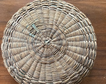 May include: A round, woven basket made of natural materials. The basket has a circular shape and a woven pattern.