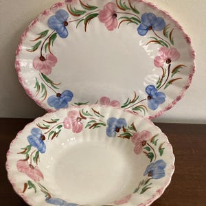 May include: A two-piece ceramic serving set comprising an oval platter and a round bowl. Both items are white with a pink, scalloped edge and decorated with pink and blue floral patterns and green leaves.