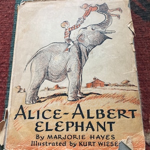 Op de afbeelding: Een vintage kinderboekomslag met een grijze olifant waarop twee kinderen rijden. De titel van het boek is "Alice-Albert Elephant" door Marjorie Hayes, geïllustreerd door Kurt Wiese.