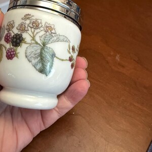 May include: A white porcelain egg coddler with a silver-colored metal lid and a ring handle. The body features a botanical illustration of green leaves, white flowers, and clusters of blackberries in shades of purple and black against a white background.