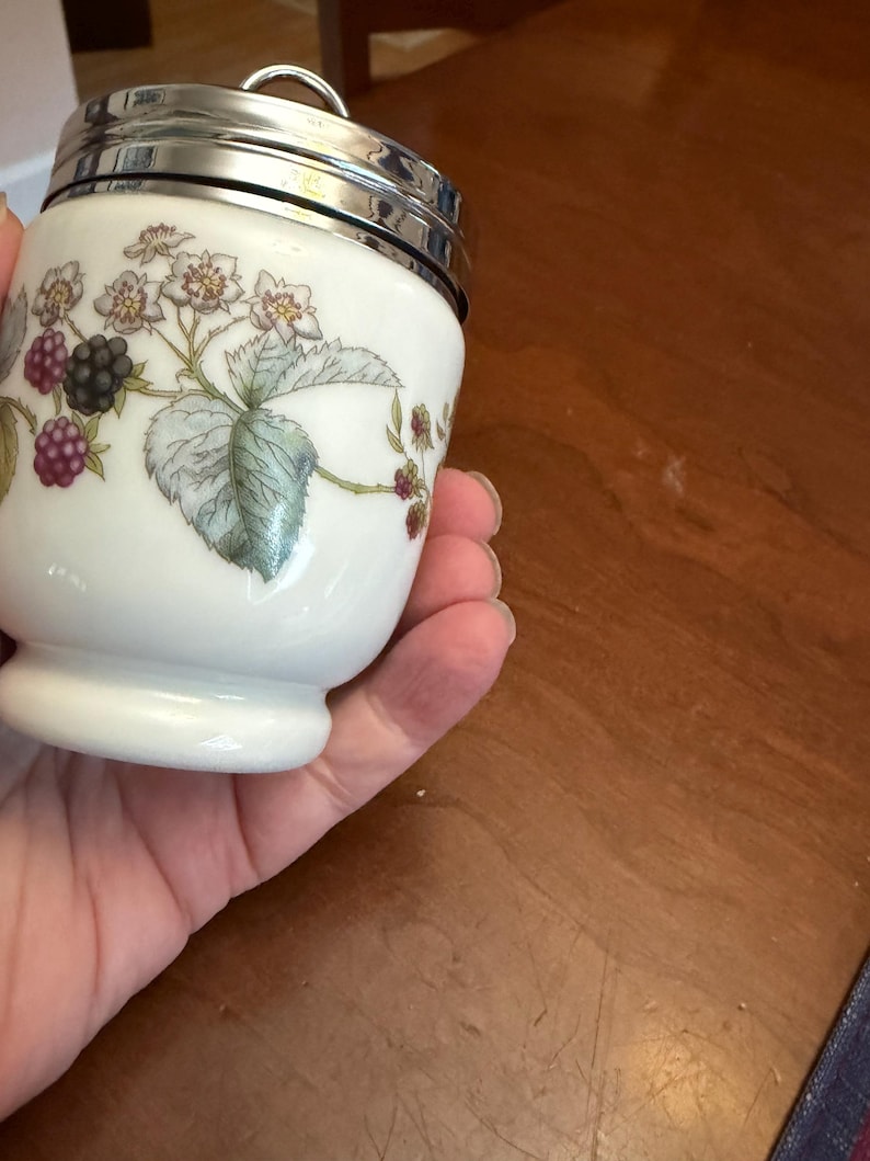 May include: A white porcelain egg coddler with a silver-colored metal lid and a ring handle. The body features a botanical illustration of green leaves, white flowers, and clusters of blackberries in shades of purple and black against a white background.
