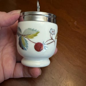 May include: A white porcelain egg coddler with a silver-colored metal lid and a ring handle. The body features a botanical pattern of green leaves, a small blue flower, and a red strawberry. The vessel has a rounded base and a smooth finish.