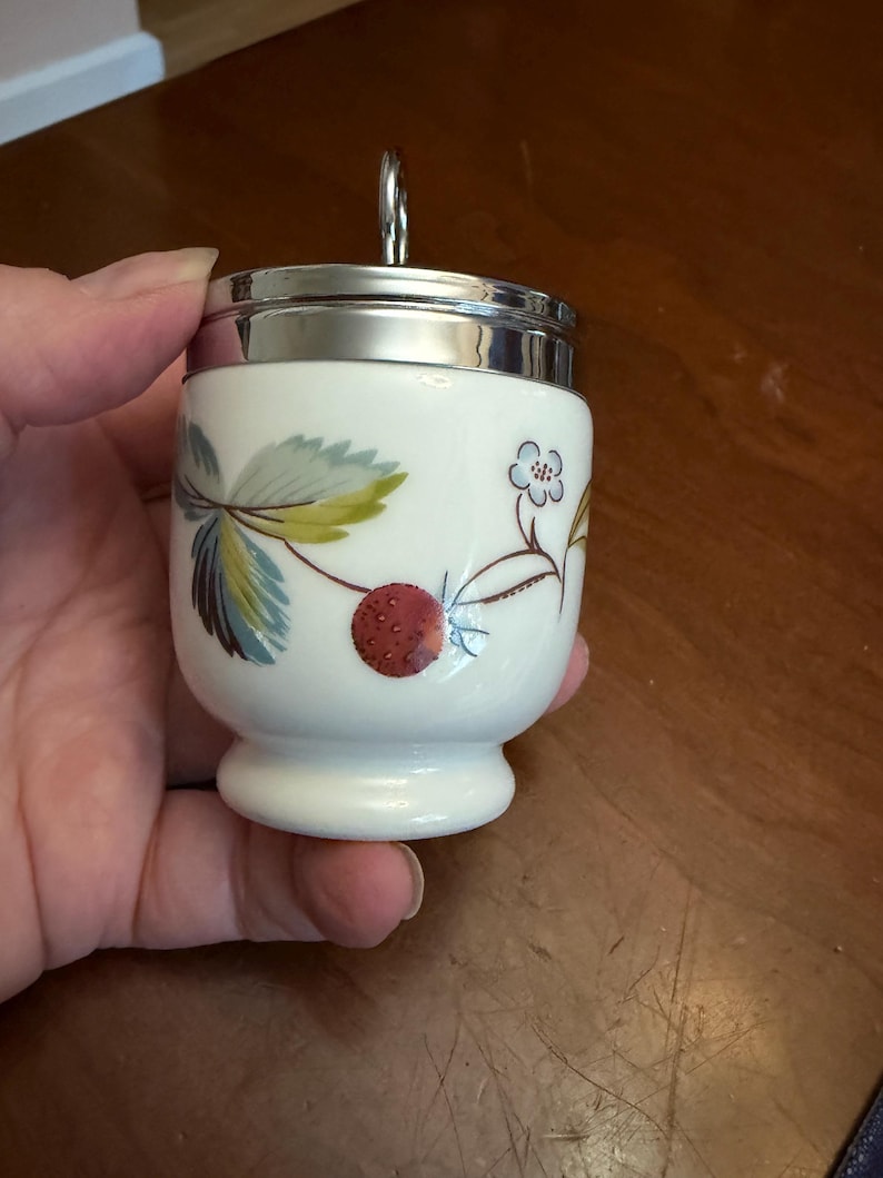 May include: A white porcelain egg coddler with a silver-colored metal lid and a ring handle. The body features a botanical pattern of green leaves, a small blue flower, and a red strawberry. The vessel has a rounded base and a smooth finish.
