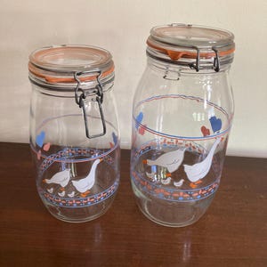 May include: Two clear glass jars with orange-rimmed lids and metal clasps. The jars are decorated with a blue, red, and white goose design. The jars are different sizes and sit on a wooden surface.