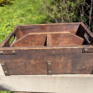 May include: A weathered, rectangular wooden box with a dark brown finish. The box features a central divider and side handles. Metal brackets and rivets reinforce the corners. The box is likely used for storage or organization.