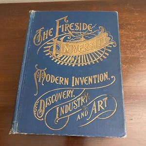 Op de afbeelding: Een blauw hardback boek met gouden letters op de omslag. De titel is "The Fireside University of Modern Invention, Discovery, Industry and Art".