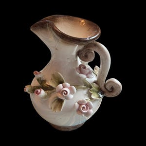 May include: A decorative ceramic pitcher with a white body, adorned with pink roses and green leaves. The rim and handle are brown. The pitcher has a curved handle and a spout for pouring.