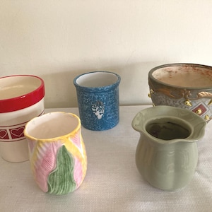 May include: Five ceramic planters in various colors and designs. One planter is white with red hearts, one is blue with a white floral design, one is green, one is yellow with pink and green leaves, and one is brown with gold accents.