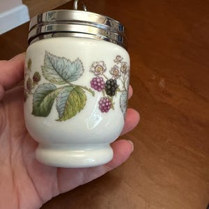 May include: A white porcelain egg coddler with a shiny silver metal lid and a small ring handle. The body features a botanical illustration of green leaves, white flowers, and clusters of purple and black blackberries encircling the container.