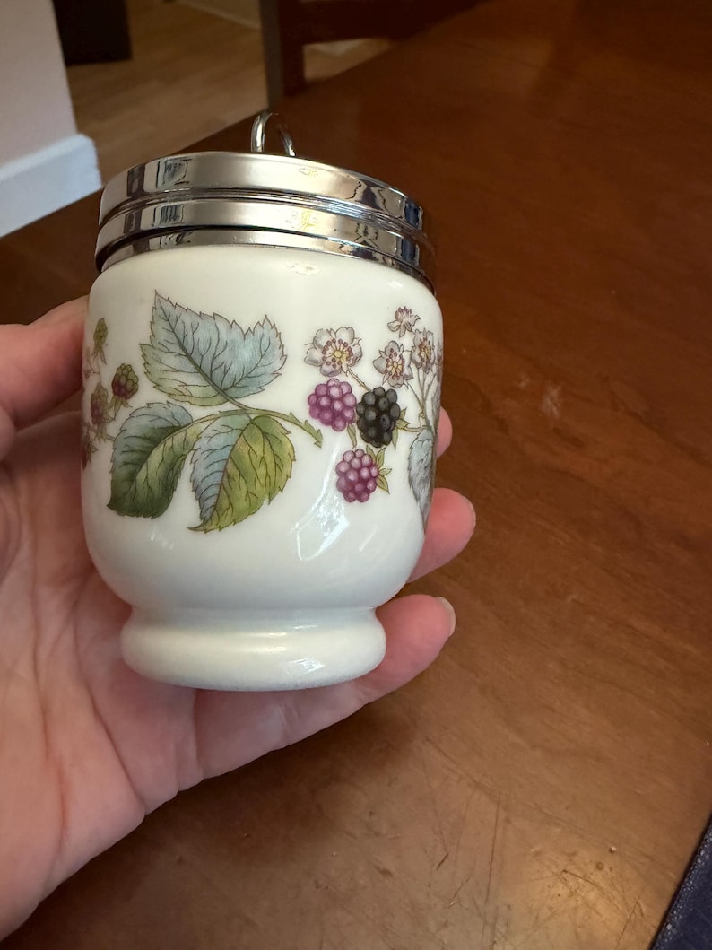 May include: A white porcelain egg coddler with a shiny silver metal lid and a small ring handle. The body features a botanical illustration of green leaves, white flowers, and clusters of purple and black blackberries encircling the container.
